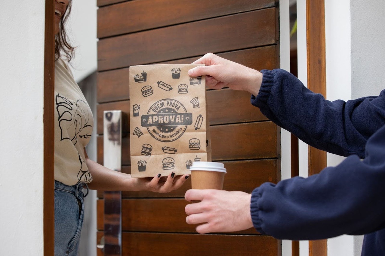 A porta está aberta e um homem entrega uma embalagem de delivery de saco de papel juntamente com um copo de papel para uma mulher.