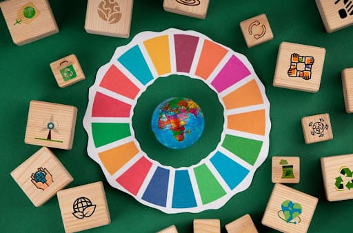 Vista de cima de uma montagem da logo do Programa ODS da ONU. Um círculo composto por cores diversas e blocos de madeira com símbolos relacionados aos ODS.