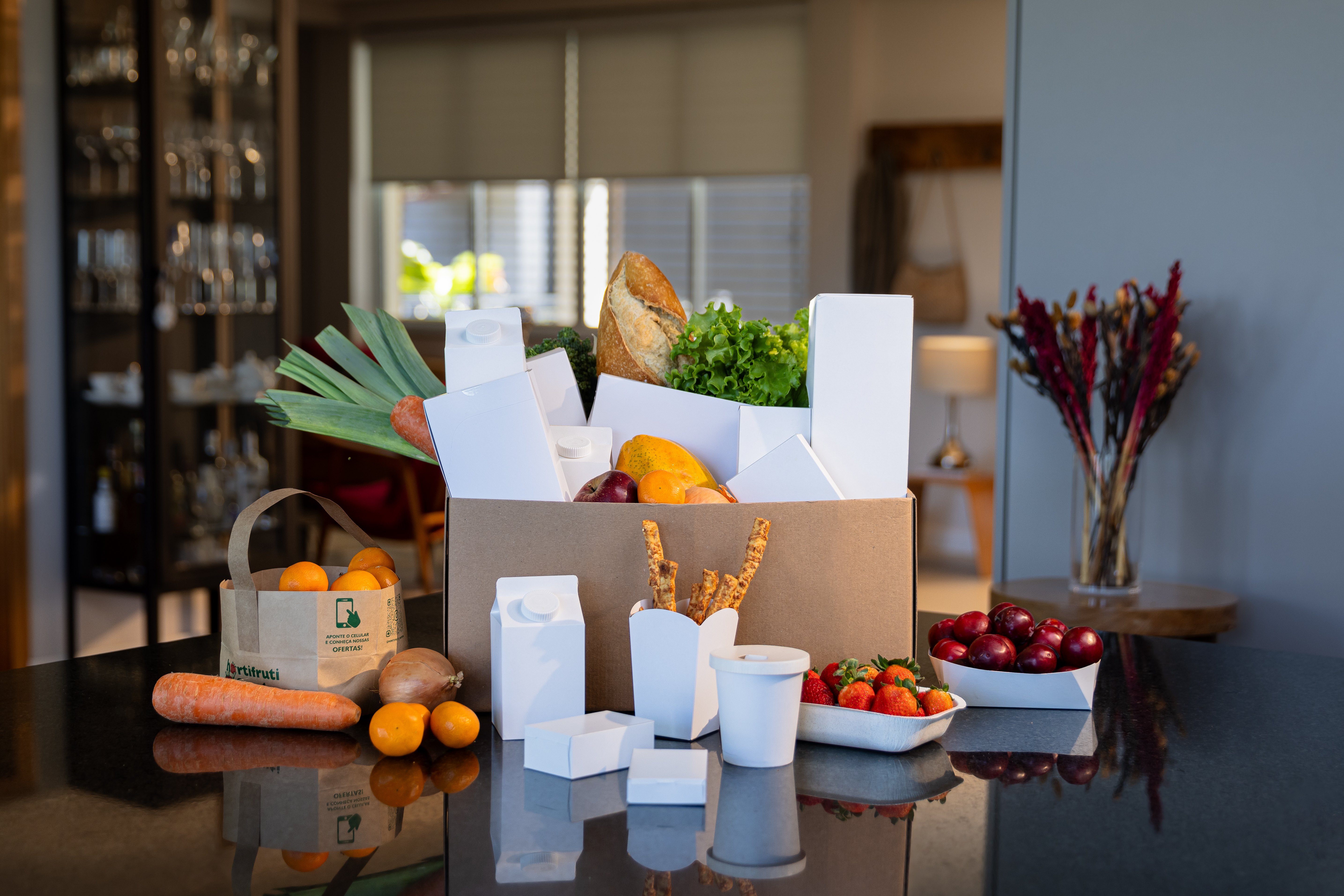 Caixas, copos e potes de papel cartão Advance sobre a mesa de uma sala, com itens como frutas e pães sendo suportados pelas emabalagens 